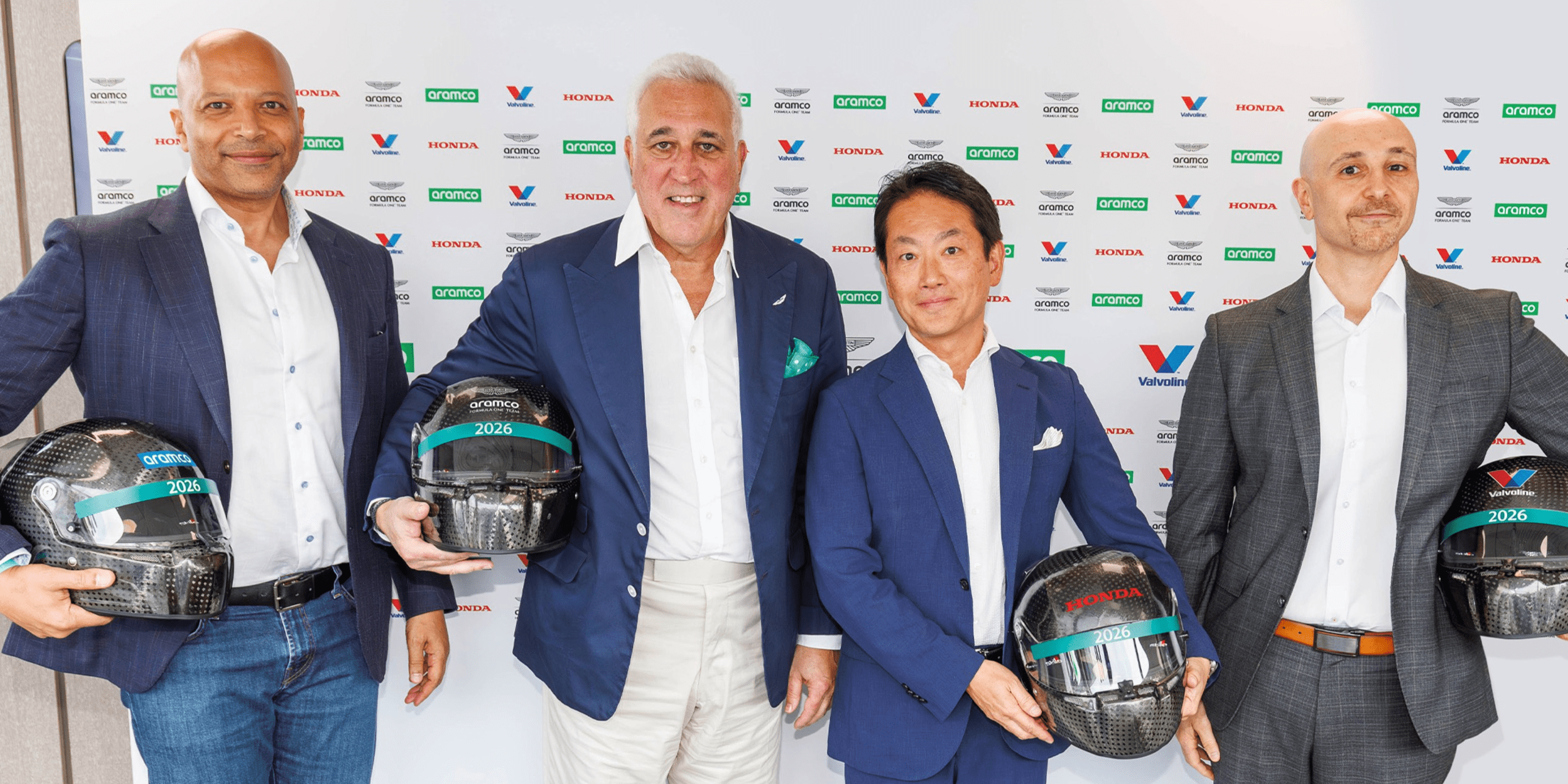 Four men wearing suit jackets and smiling, holding race helmets, in front of a sponsored backdrop