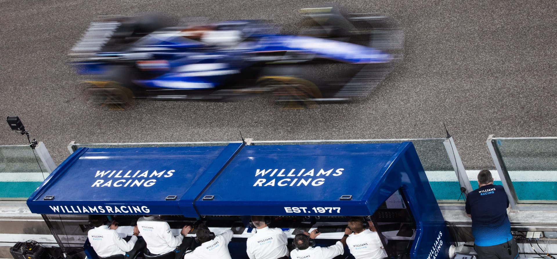 Blue F1 car, blurry, going at high speed past the Williams Racing pitwall 